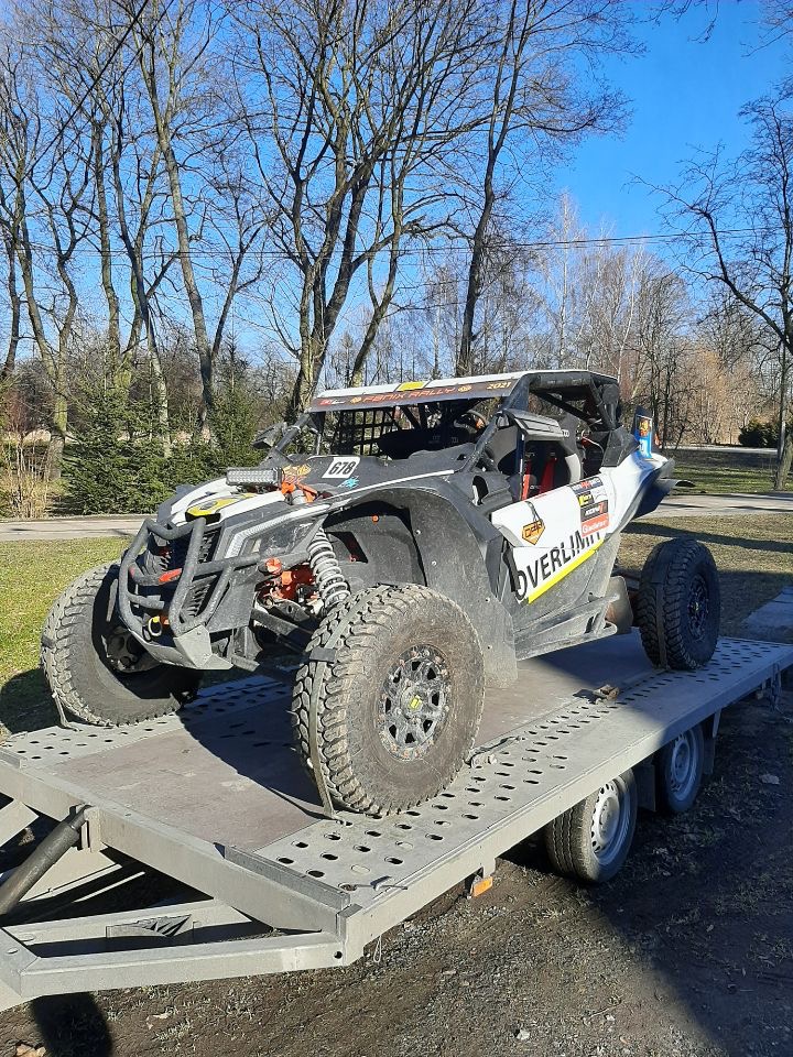 Biało-czarny pojazd terenowy typu buggy z numerem 678 na przyczepie, duże opony terenowe, widoczne logo 'OVERLIMIT', tło z drzewami bez liści i błękitnym niebem.