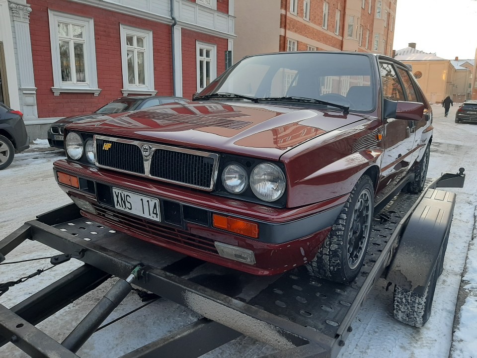 Czerwony samochód marki Lancia Delta HF Integrale z numerem rejestracyjnym XNS 114 przewożony na niskopodwoziowej przyczepie samochodowej na tle miejskiej zabudowy.