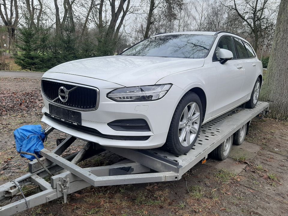 Białe Volvo kombi na metalowej przyczepie samochodowej z niebieskim pokrowcem zabezpieczającym na haku holowniczym, tło park z drzewami.