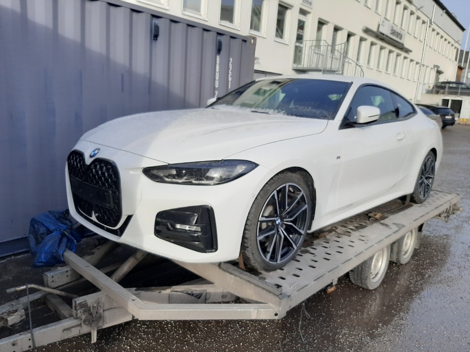 Białe BMW serii 4 na niskopodwoziowej przyczepie z widocznymi śladami użytkowania, w tle budynek z białą elewacją i kontener.