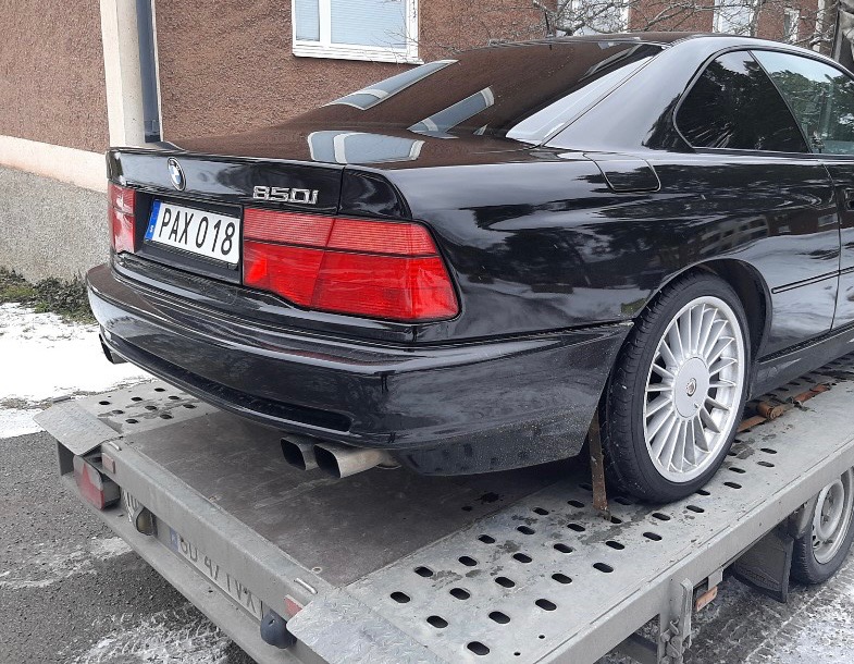 Czarny samochód BMW 850i z tablicą rejestracyjną PAX 018, widoczny z tyłu, umieszczony na platformie transportowej z metalowymi otworami, prawdopodobnie po awarii.