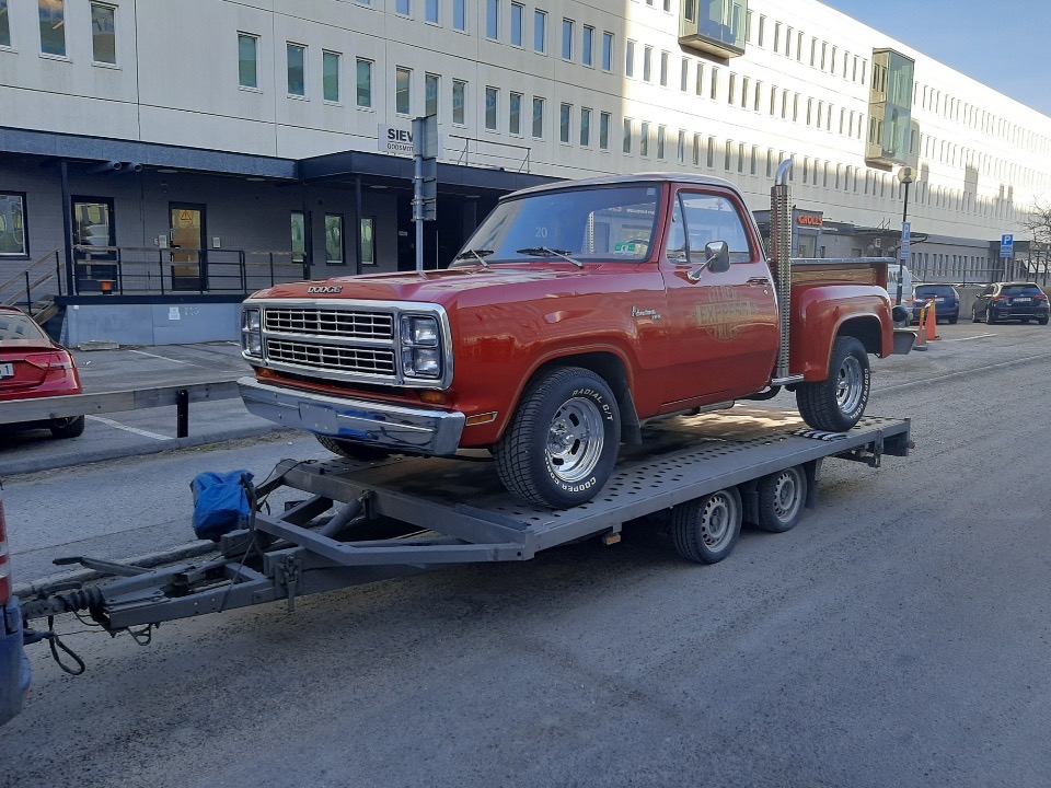 Czerwony Dodge Ram Express Truck z lat 70. transportowany na niskopodwoziowej przyczepie lawetowej, widok z przodu, budynek biurowy w tle.