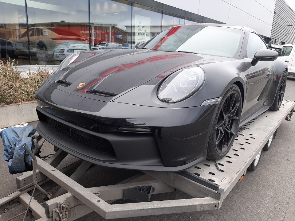 Czarny Porsche 911 na niskopodwoziowej lawecie transportowej przed salonem samochodowym.