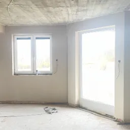 Wnętrze w stanie deweloperskim: ściany pokryte świeżym tynkiem, surowy betonowy sufit z widocznymi instalacjami, okno i drzwi balkonowe w białych ramach, wystające przewody elektryczne.
