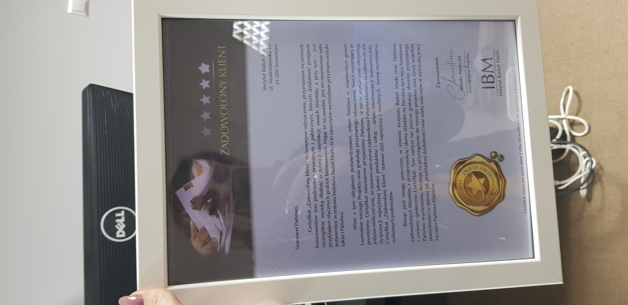 Oprawiony certyfikat 'Zadowolony Klient' Instytutu Badań Marki, widoczny obok monitora Dell, prezentuje wyróżnienie za dbałość o klienta i etyczne praktyki biznesowe.
