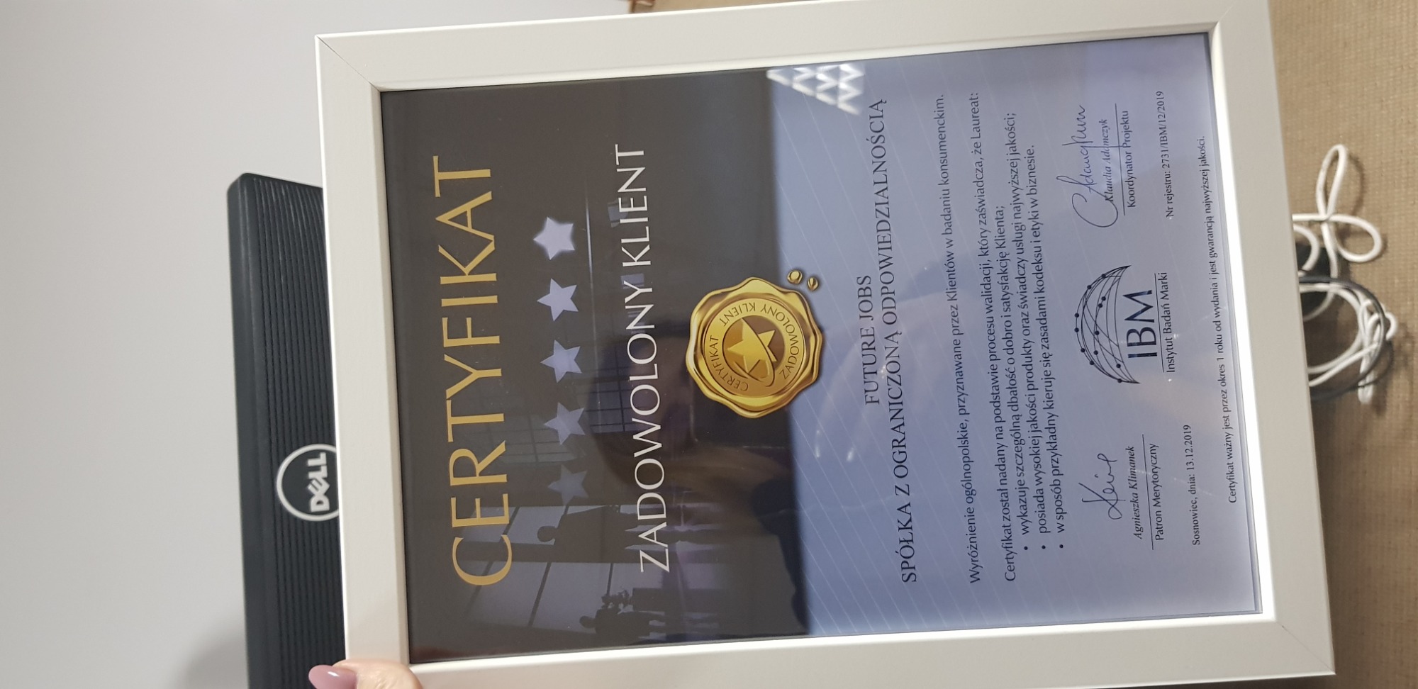 Oprawiony w białą ramkę certyfikat 'Zadowolony Klient' dla Future Jobs Spółka z ograniczoną odpowiedzialnością, przyznany przez Instytut Badań Marki IBM, leżący obok czarnego laptopa Dell.