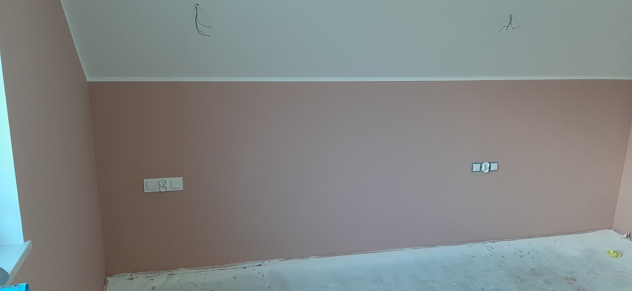 Ściana pomalowana na różowo, z zamontowanymi gniazdkami elektrycznymi i wystającymi przewodami z sufitu, podłoga zabezpieczona folią malarską.