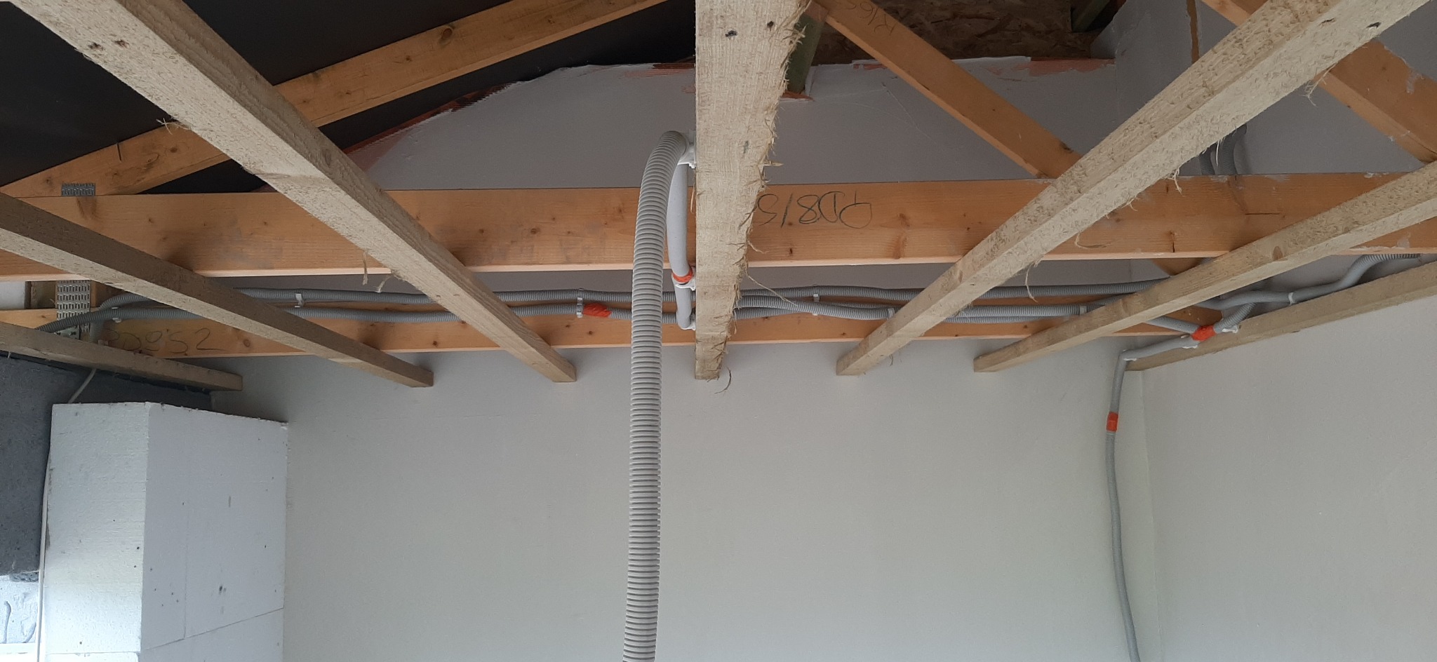 Szare rury instalacji elektrycznej zamocowane do drewnianych belek stropowych w surowym stanie, widoczne napisy na belce i fragment ściany z białym styropianem.