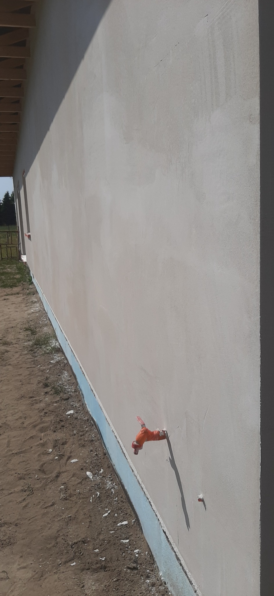 Świeżo otynkowana elewacja budynku z widocznym kranem zewnętrznym z tworzywa sztucznego w kolorze pomarańczowym, osadzonym w ścianie. Widoczne ślady po pracach wykończeniowych i niedoskonałości tynku.