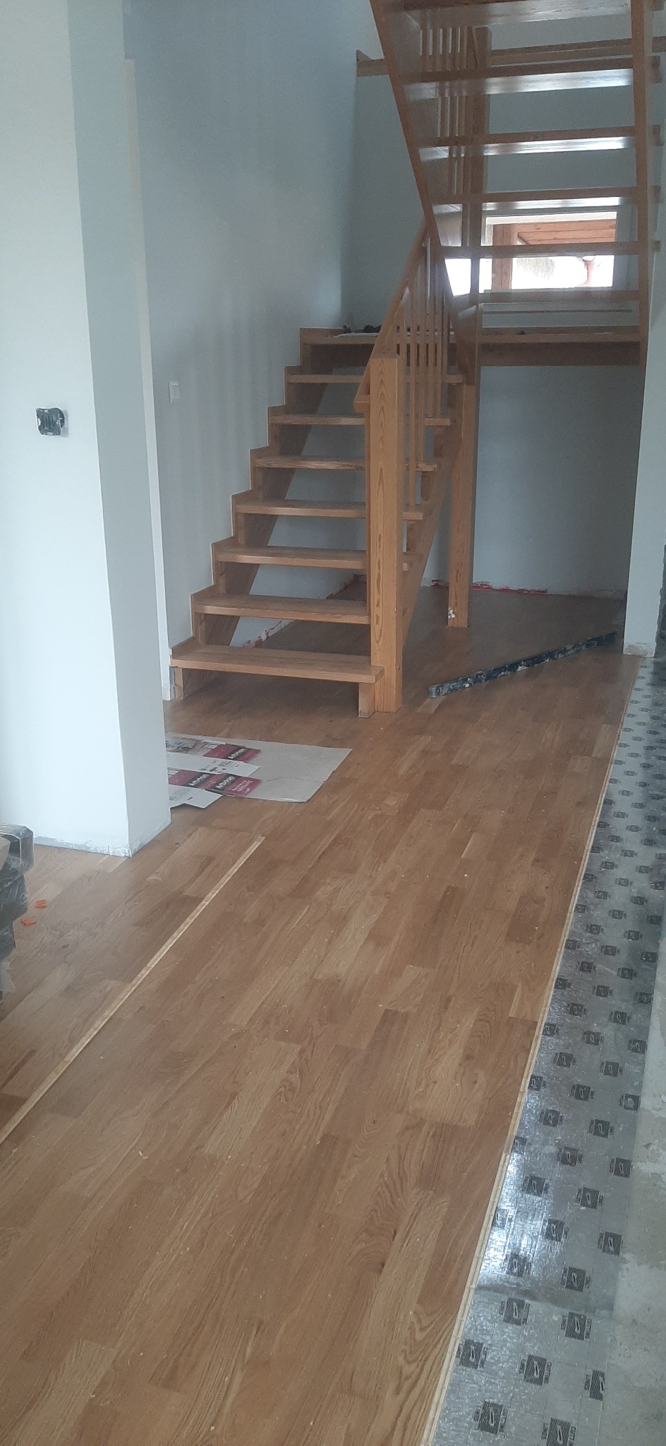 Układanie drewnianej podłogi w jasnym odcieniu, widoczne schody drewniane prowadzące na górę, częściowo położona podłoga z widoczną folią izolacyjną na podłożu.