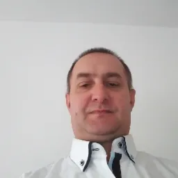 Portret mężczyzny w białej koszuli z czarnymi akcentami pod szyją, na jasnym tle. Ujęcie z dołu, widoczna część głowy i ramion.