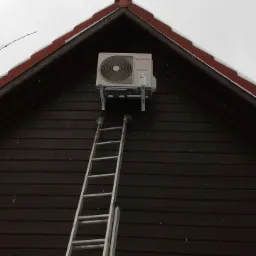 Zewnętrzna jednostka klimatyzacji Toshiba zamontowana na szczycie drewnianego domu z użyciem metalowych wsporników, z postawioną obok aluminiową drabiną, widok z dołu.