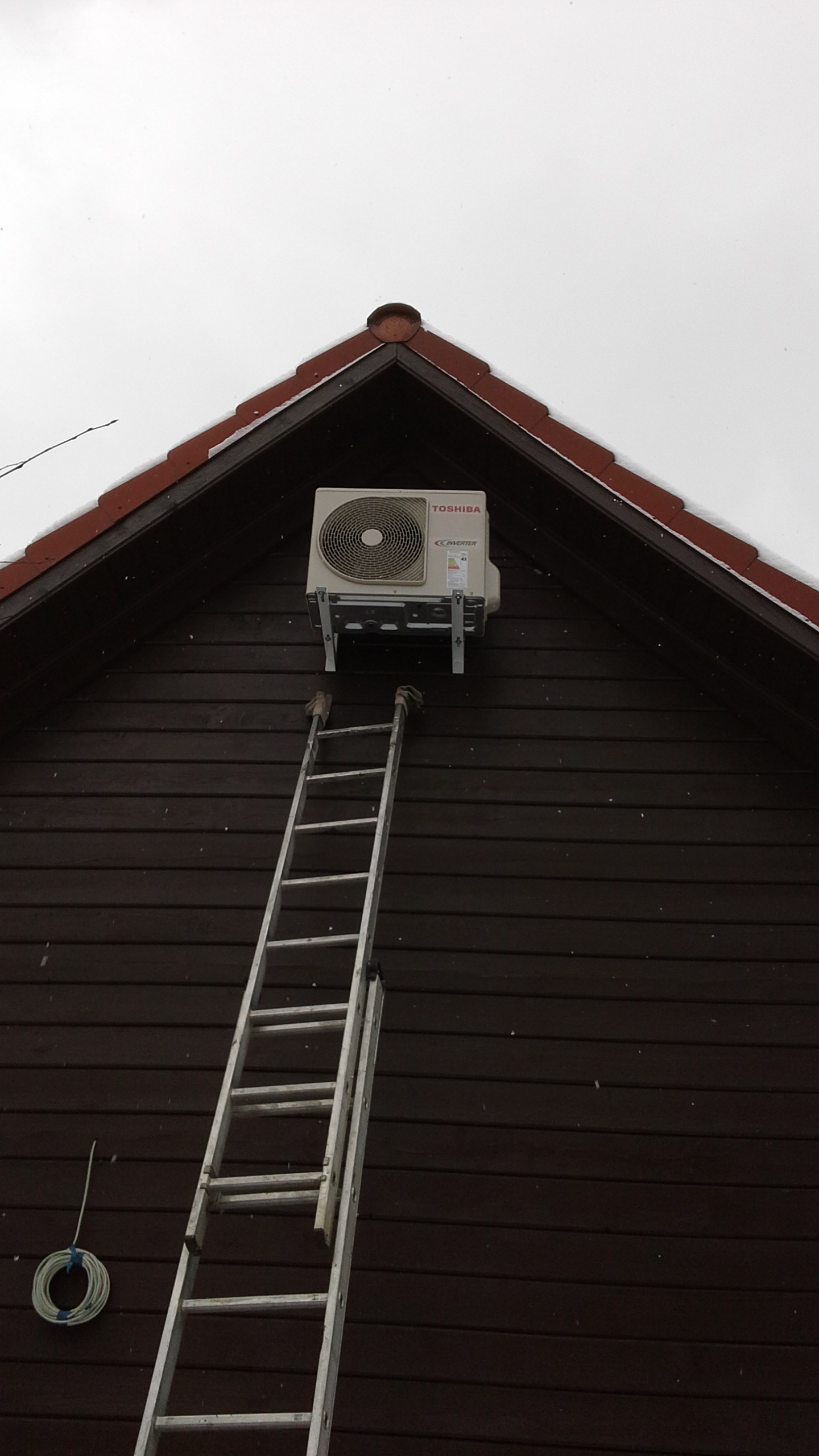 Zewnętrzna jednostka klimatyzacji Toshiba zamontowana na szczycie drewnianego domu z użyciem metalowych wsporników, z postawioną obok aluminiową drabiną, widok z dołu.