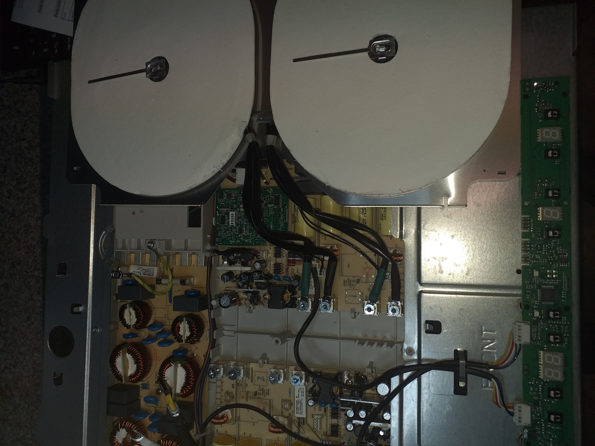 Wnętrze naprawianej płyty indukcyjnej z widocznymi cewkami, elektroniką i elementami grzejnymi z białymi osłonami, naprawa w toku.