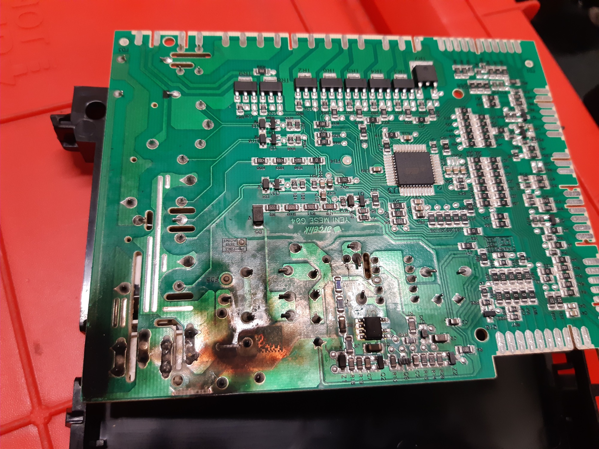 Uszkodzona płyta sterująca urządzenia AGD z widocznymi śladami przypalenia, umieszczona na czerwonym podłożu.