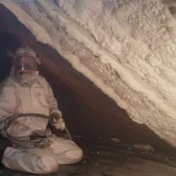 Pracownik w kombinezonie ochronnym klęczy w ciemnym poddaszu, trzymając wąż natryskowy, obok świeżo nałożonej, grubej warstwy pianki PUR na skośnym dachu.