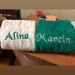 Dwa złożone, puszyste ręczniki: biały z wyhaftowanym zielonym napisem 'Alina' i zielony z wyhaftowanym białym napisem 'Marcin', leżące na drewnianej komodzie, w tle świeczka i mała roślina.