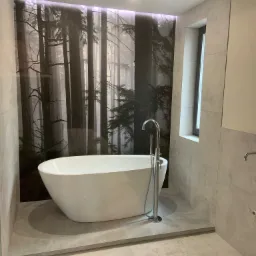 Wolnostojąca wanna z chromowaną baterią na tle fototapety z motywem leśnym, szare płytki na ścianach i podłodze, oświetlenie punktowe w suficie.