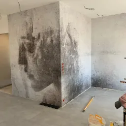 Wnętrze pokoju w trakcie prac wykończeniowych, ściany pokryte tapetą imitującą beton z abstrakcyjnym motywem twarzy, widoczne przewody elektryczne wystające z sufitu, wiadro z klejem i narzędzia...