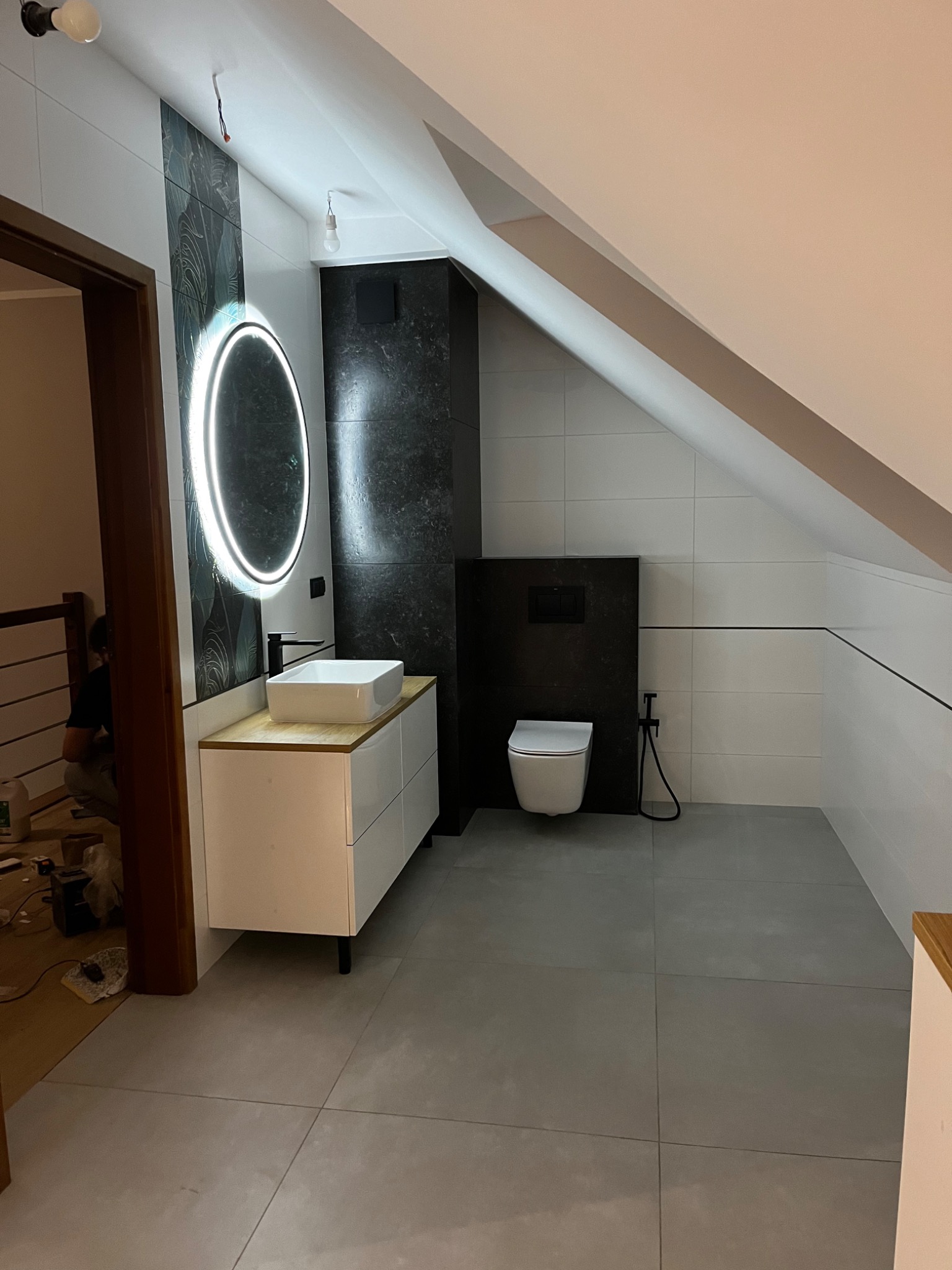 Wykończona łazienka na poddaszu z szarą podłogą, białą armaturą, umywalką nablatową na białej szafce z blatem imitującym drewno oraz okrągłym lustrem z podświetleniem LED. Widoczne białe płytki...