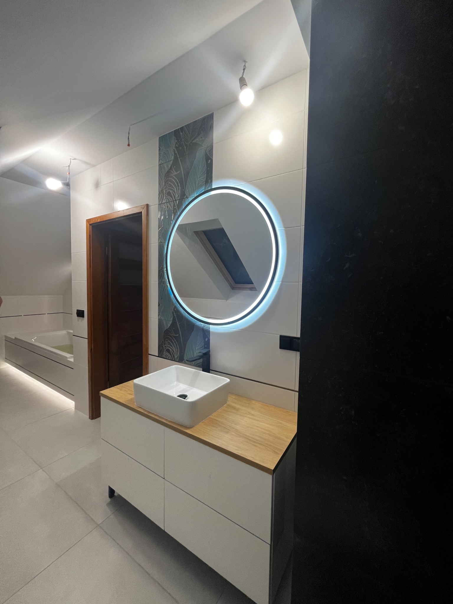 Nowoczesna łazienka z podświetlanym okrągłym lustrem, białą umywalką nablatową na szafce z drewnianym blatem, i płytkami z motywem liści na ścianie.