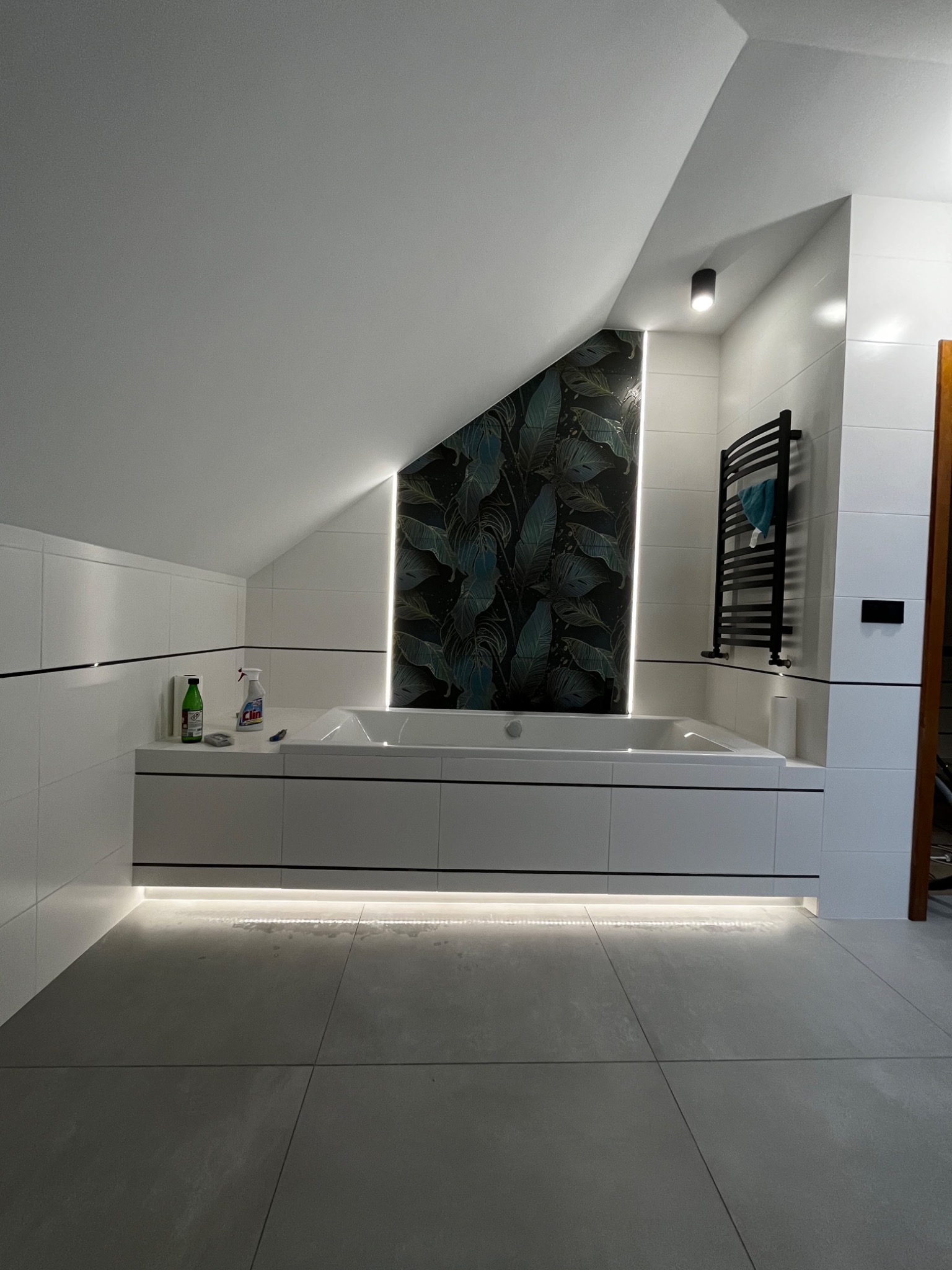 Nowoczesna łazienka na poddaszu z białą zabudową wanny, podświetleniem LED pod szafkami i pionowym panelem dekoracyjnym z motywem roślinnym, widoczny grzejnik drabinkowy i butelki środków czystości.