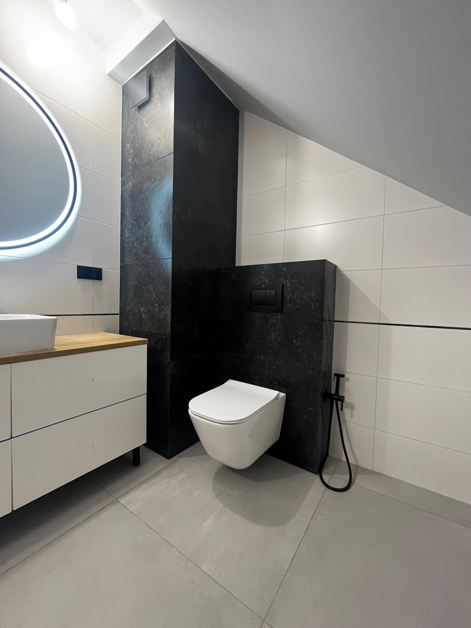 Nowoczesna łazienka na poddaszu z biało-czarną kolorystyką, podwieszana toaleta z czarną armaturą bidetową, biała szafka pod umywalką z blatem z jasnego drewna, okrągłe lustro z podświetleniem LED.