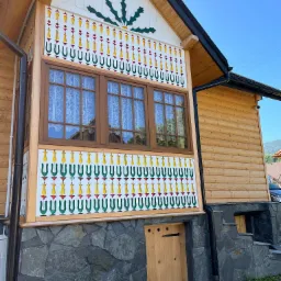 Fasada domu z drewnianą elewacją, zdobiona tradycyjnymi, kolorowymi wzorami ludowymi, okna z białymi koronkowymi firankami, kamienna podmur&oacute;wka z małym drewnianym wejściem.