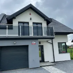 Nowoczesny dom dwukondygnacyjny z balkonem z balustradą ze szkła i szarą bramą garażową, widoczny znak firmy ochroniarskiej w oknie.