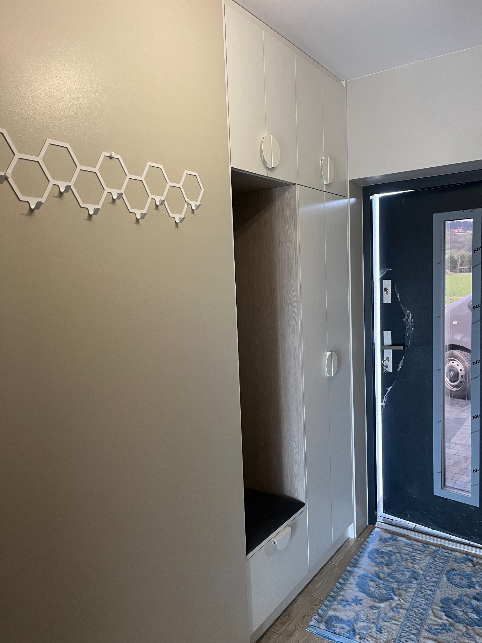 Nowoczesny przedpokój z białą zabudową szaf, siedziskiem i wieszakiem w kształcie sześciokątów na ścianie, widok na zewnątrz przez otwarte drzwi wejściowe.