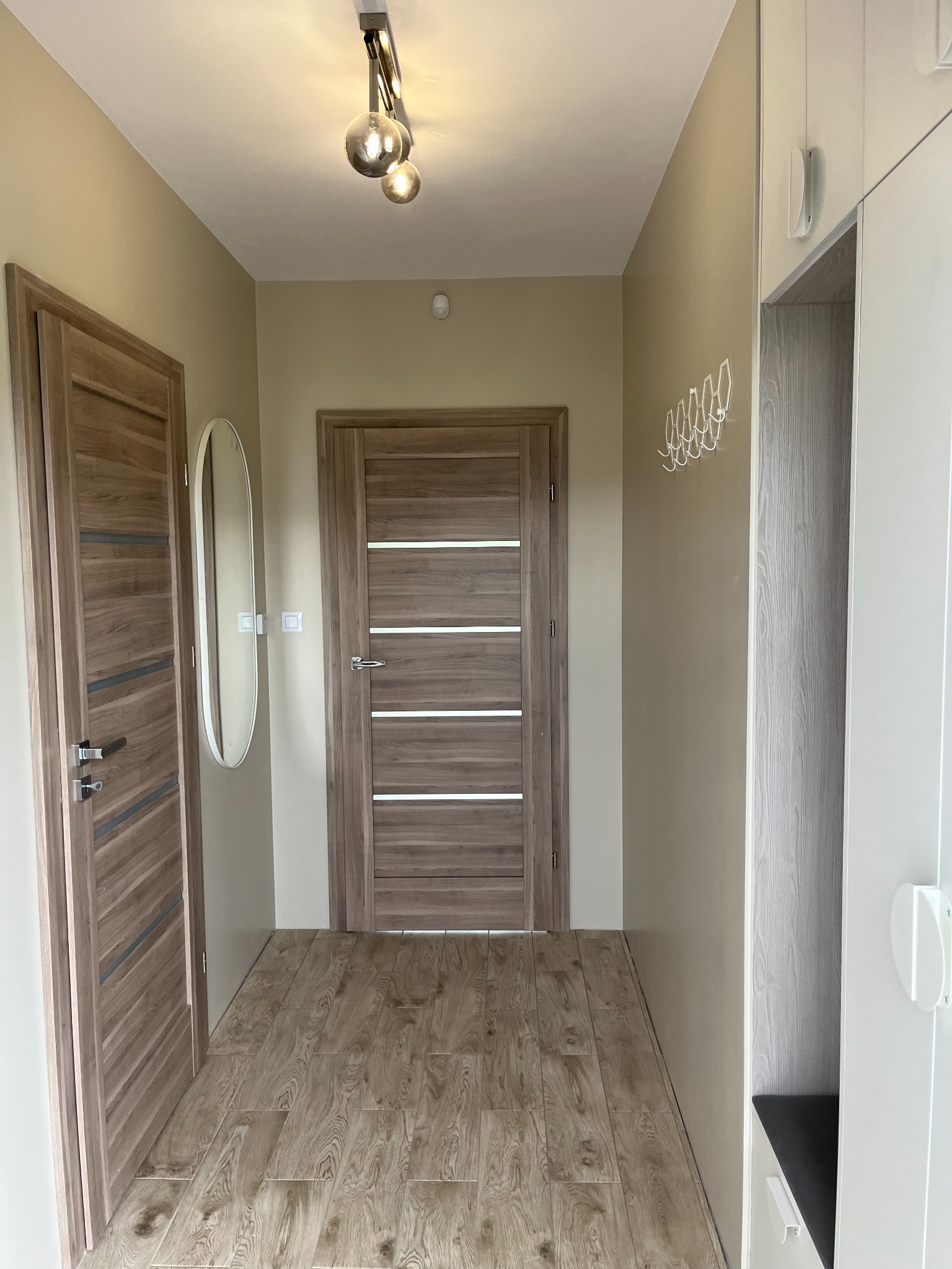 Nowoczesny przedpokój z dwiema drewnianymi drzwiami z poziomymi wstawkami, owalnym lustrem, drewnopodobną podłogą, białymi wieszakami i szafą w zabudowie z siedziskiem.