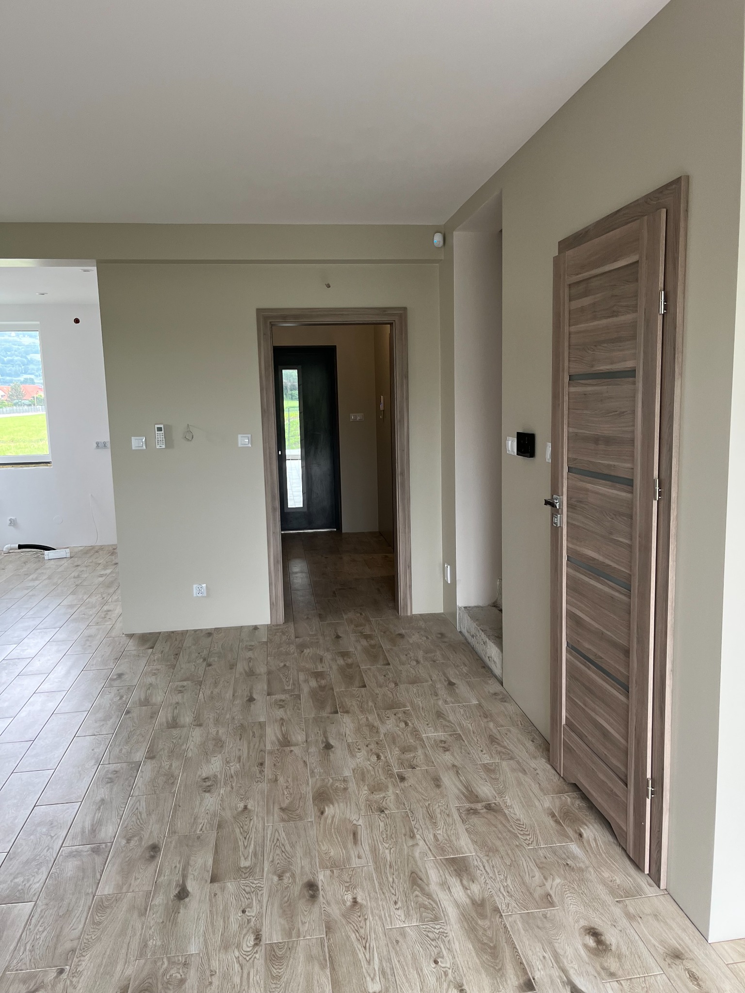 Wnętrze w trakcie wykańczania: drewnopodobna podłoga, beżowe ściany, drzwi wewnętrzne z drewnianym fornirem, widok na okno i krajobraz za nim.