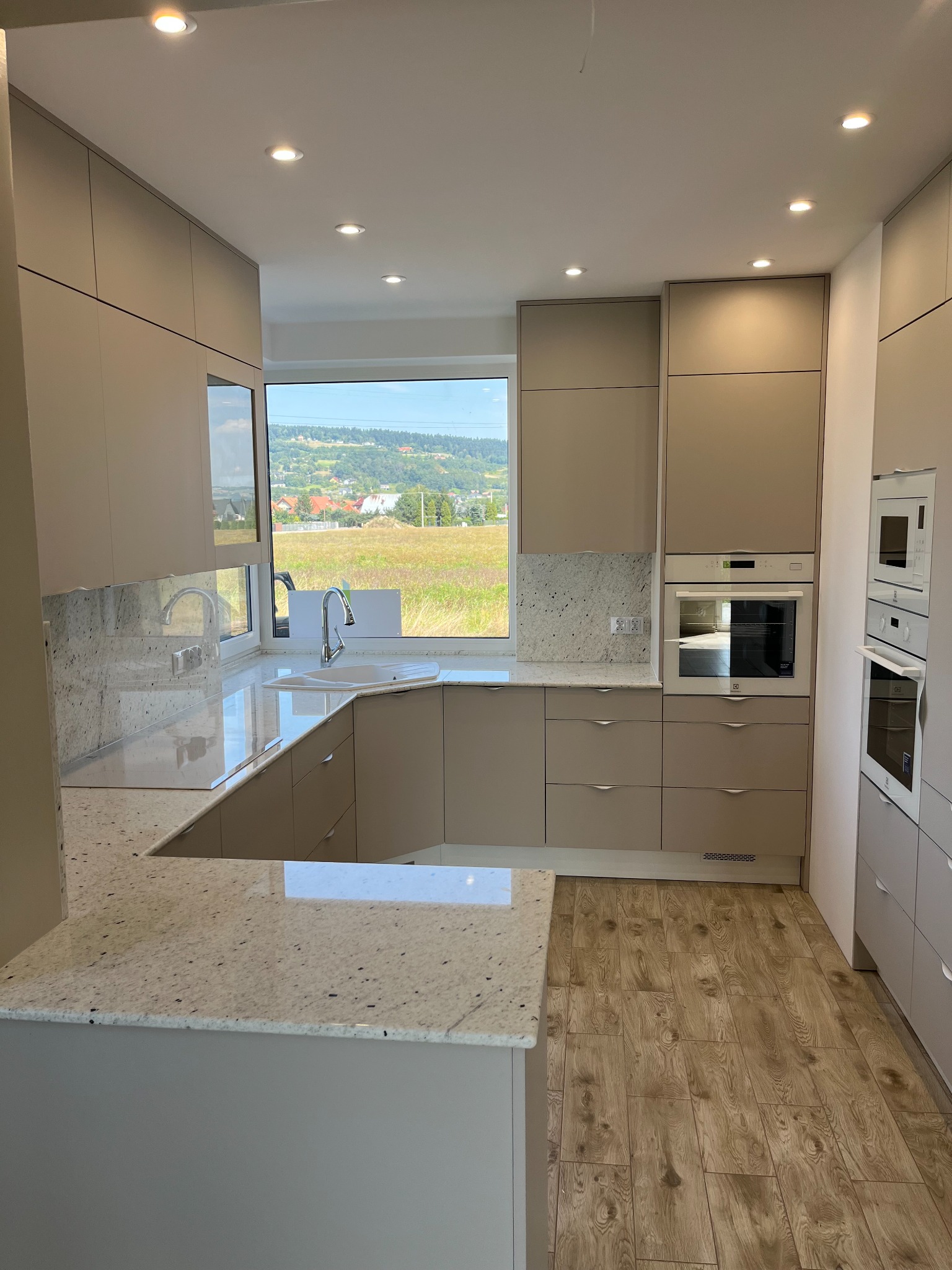Jasna kuchnia z beżowymi szafkami i blatem z jasnego granitu, widok na zielone wzgórza przez okno, drewnopodobna podłoga.