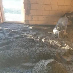 Aplikacja jastrychu cementowego na podłodze w surowym stanie; widoczne fragmenty siatki zbrojeniowej i pracownik w kombinezonie wyrównujący masę.