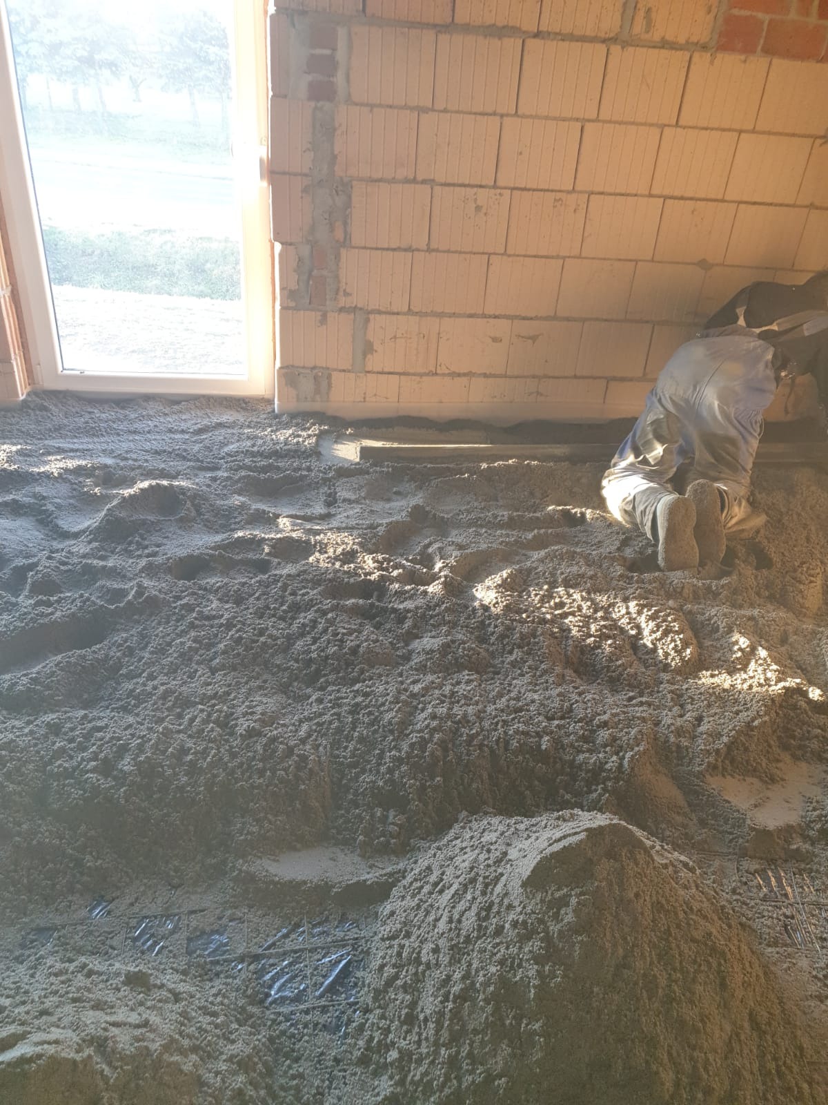 Aplikacja jastrychu cementowego na podłodze w surowym stanie; widoczne fragmenty siatki zbrojeniowej i pracownik w kombinezonie wyrównujący masę.