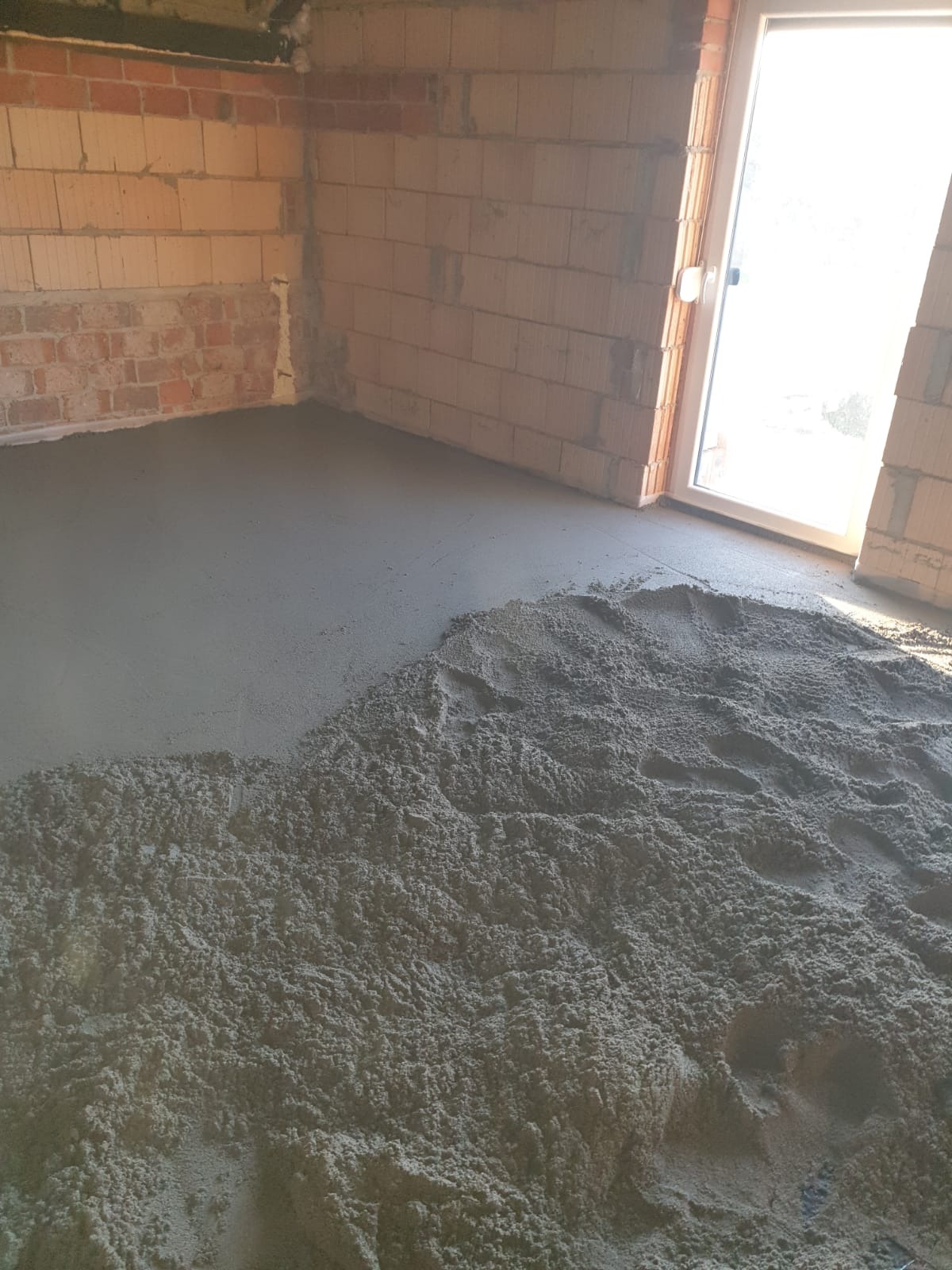 Wylewka jastrychowa w trakcie realizacji, widoczna świeżo wylana, gładka powierzchnia oraz sterta mokrego jastrychu do rozprowadzenia w pomieszczeniu z surowymi ścianami z cegły.