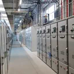 Długi korytarz z szarymi szafami elektrycznymi po obu stronach, z pracownikami w kamizelkach odblaskowych pracującymi na konstrukcji pod sufitem, widoczne kable i rury pod sufitem.