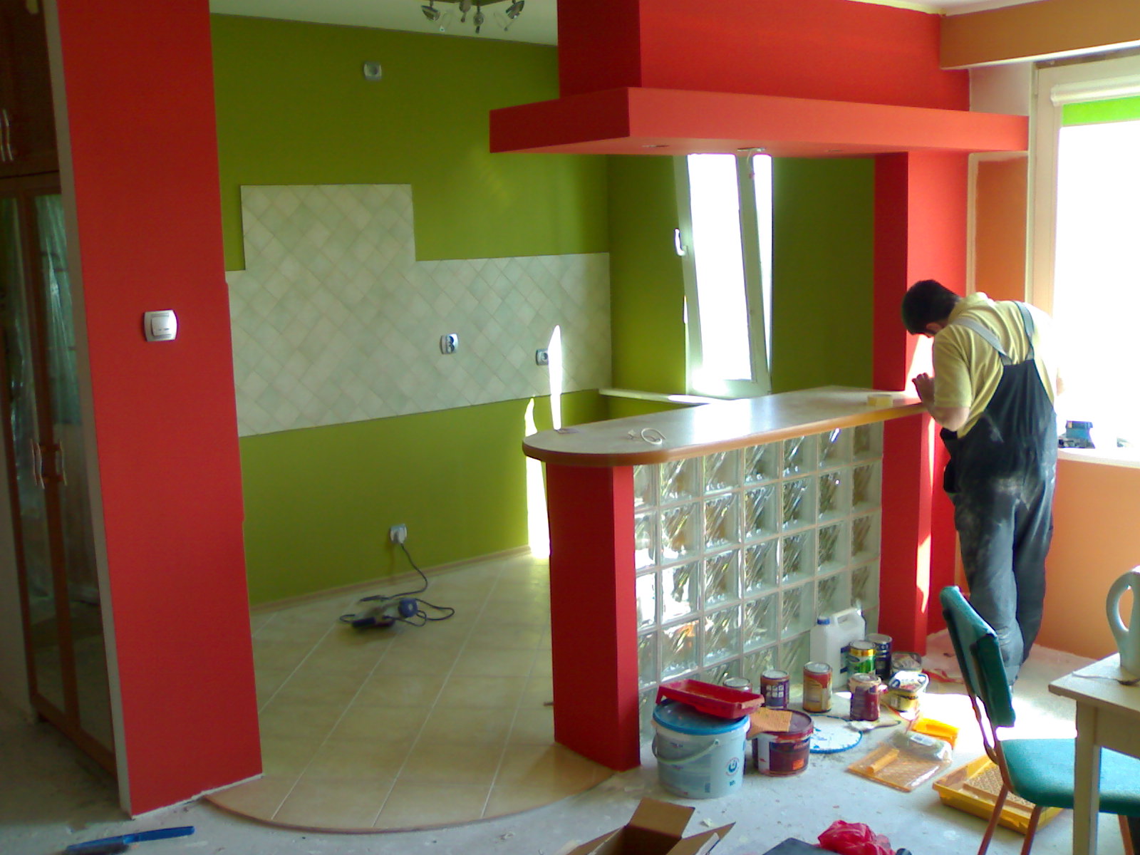 Wnętrze w trakcie malowania: ściany w odcieniach zieleni i czerwieni, bar z luksferów, widoczne puszki z farbą i narzędzia malarskie, pracownik w kombinezonie wykonujący prace wykończeniowe przy...