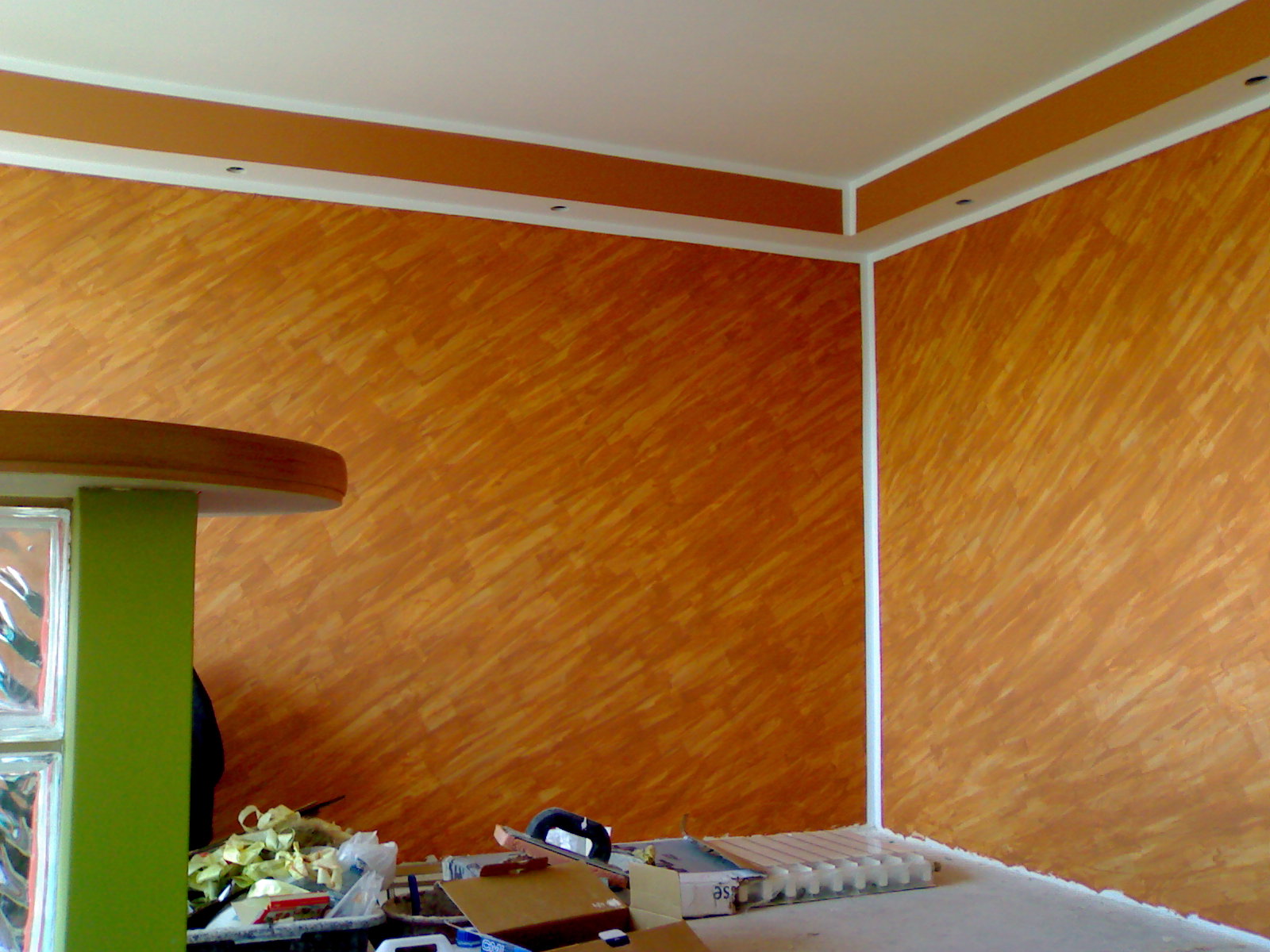 Ściana w odcieniach pomarańczy z dekoracyjnym wzorem, białe listwy przypodłogowe i sufitowe, z elementami przygotowań do prac wykończeniowych widocznymi na podłodze.