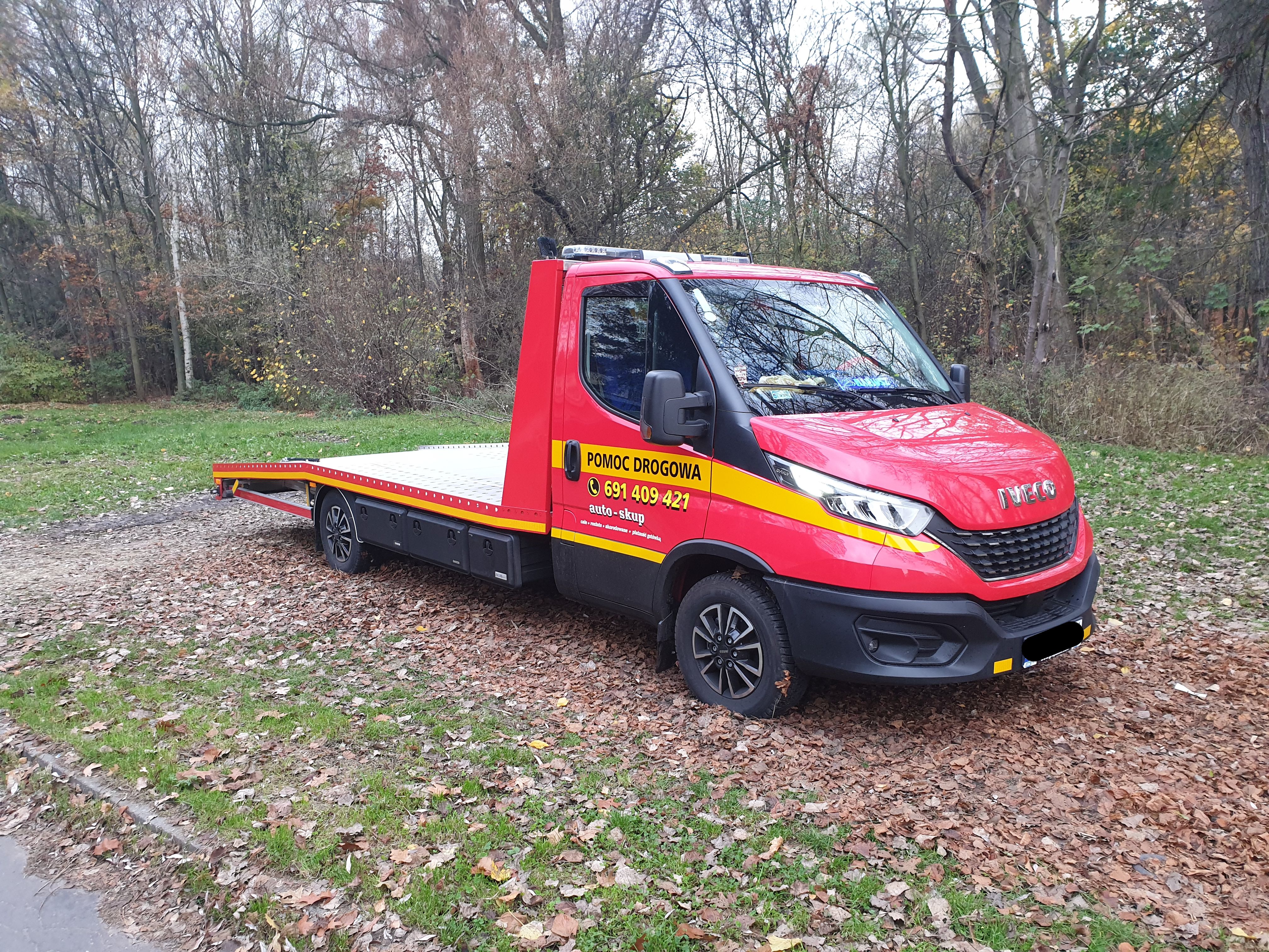 Czerwona laweta Iveco z napisem 'Pomoc Drogowa' i numerem telefonu, zaparkowana na trawniku pokrytym opadłymi liśćmi, z drzewami w tle.