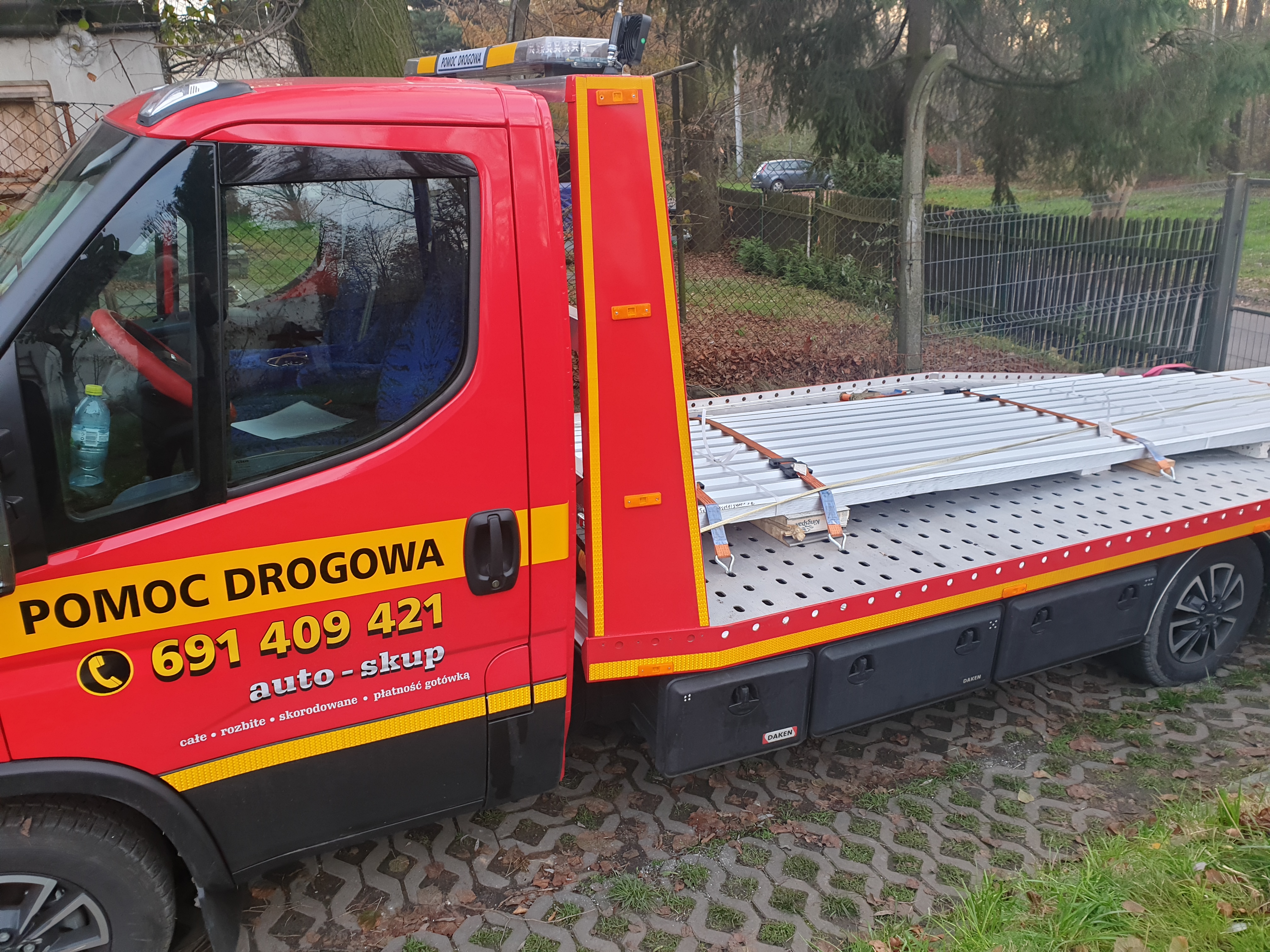 Czerwona laweta z napisem 'POMOC DROGOWA' i numerem telefonu, zaparkowana na kostce brukowej, z ładunkiem zabezpieczonym pasami na platformie.