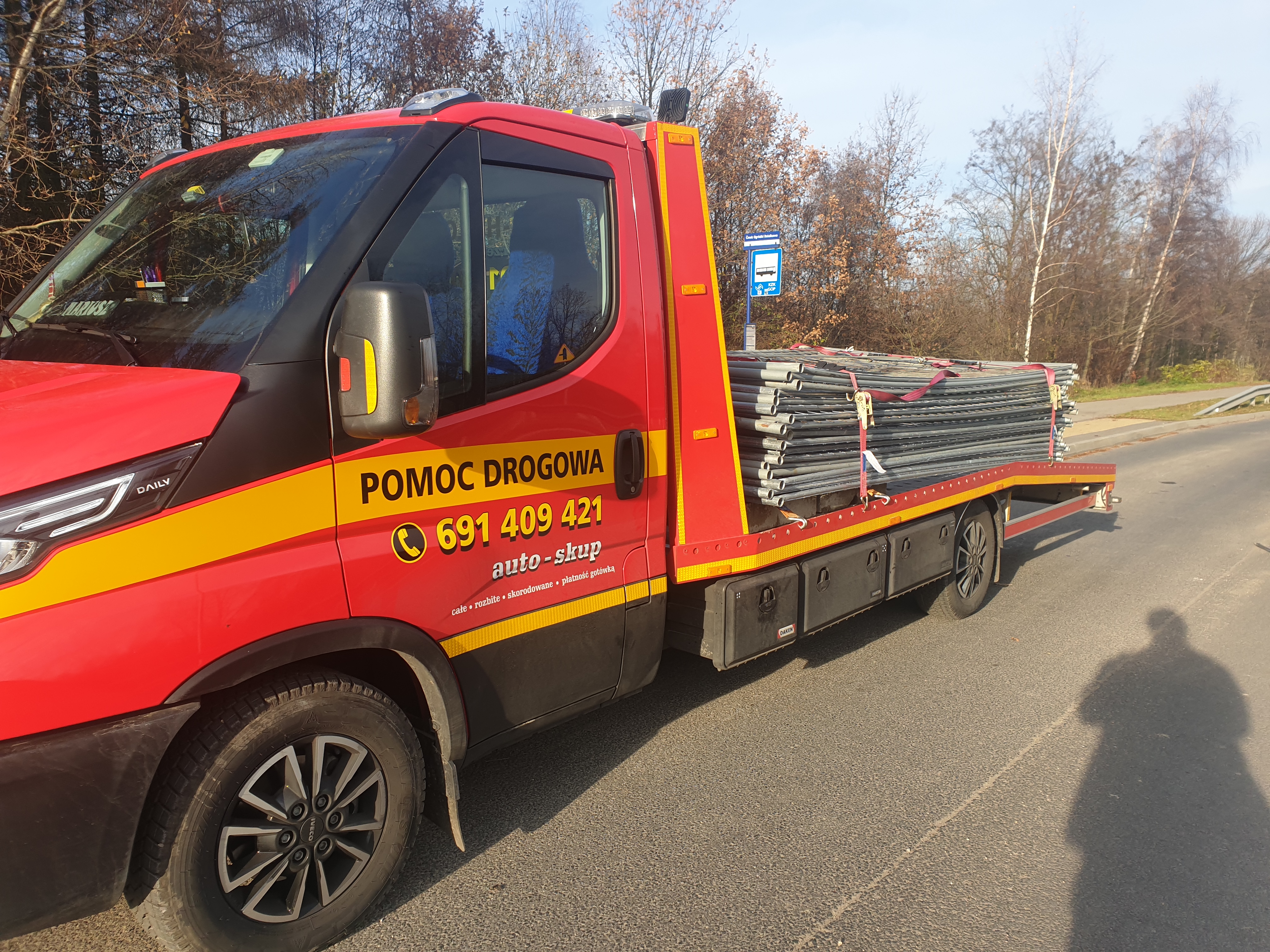 Czerwona ciężarówka z napisem 'Pomoc Drogowa' przewozi zabezpieczony ładunek metalowych rur na platformie, stojąc na asfalcie przy drodze w słoneczny dzień.