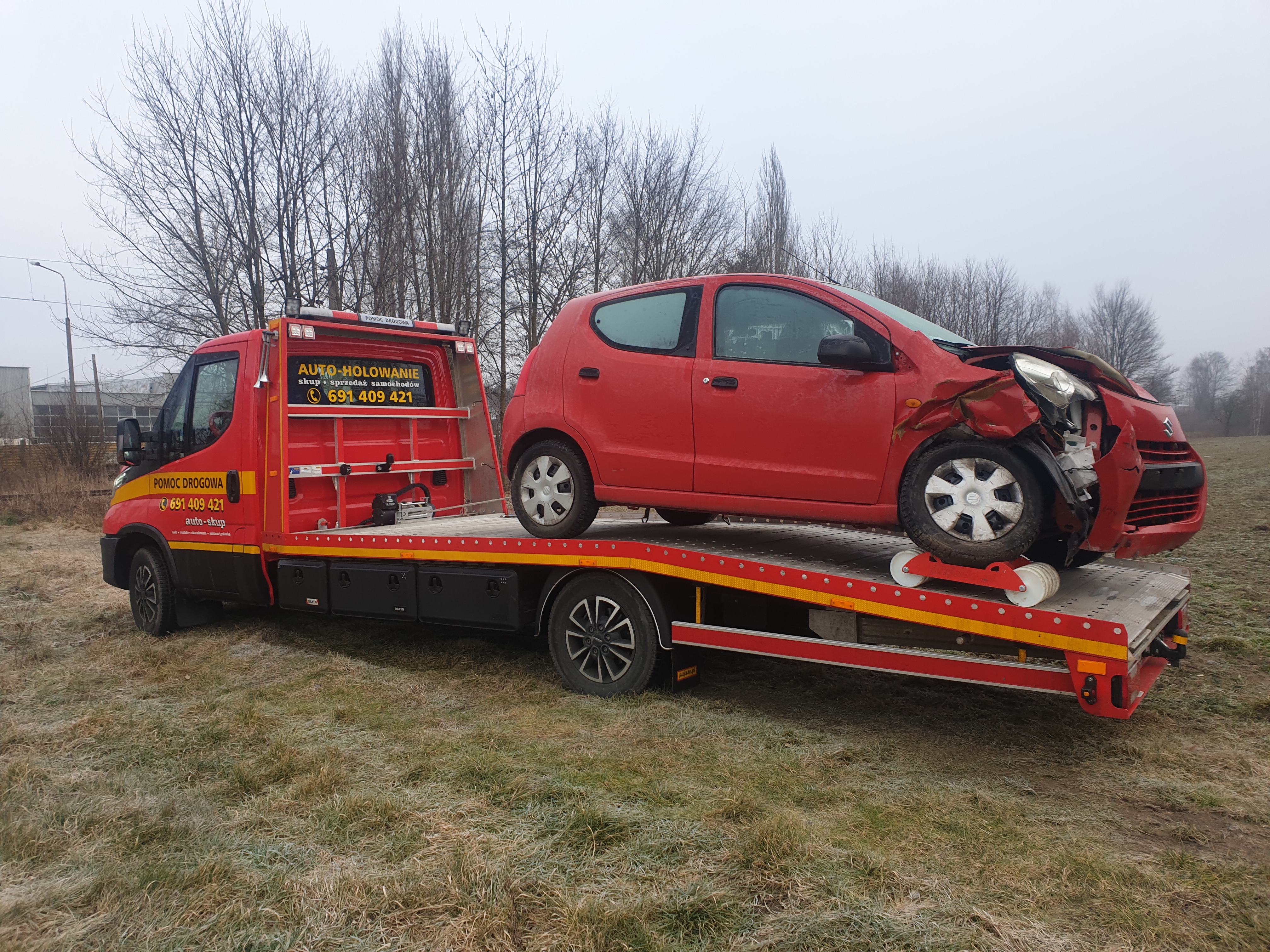 Czerwony uszkodzony samochód osobowy na platformie czerwono-czarnej lawety z napisem 'AUTO-HOLOWANIE skup sprzedaż samochodów 691 409 421' na tle drzew bez liści i zabudowań.