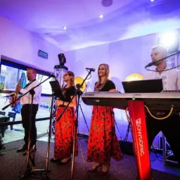 Czteroosobowy zespół muzyczny w strojach ludowych, grający na saksofonie, klarnecie i keyboardzie, oświetlony niebieskim światłem, z dużymi lampami w tle.