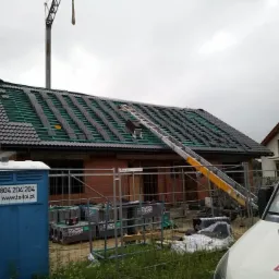 Montaż ciemnej dachówki na nowym domu z cegły, widoczne rusztowanie i toaleta przenośna, pochmurne niebo.