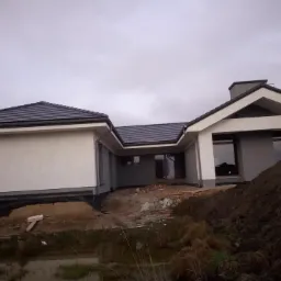 Nowoczesny dom jednorodzinny w trakcie wykończenia, widoczny fragment dachu z szarej dachówki, świeżo otynkowana elewacja w odcieniach szarości i bieli, otoczenie w stanie surowym z hałdami ziemi...