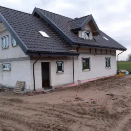 Nowo wybudowany dom jednorodzinny z czarną dachówką, oknami z folią ochronną i otwartym wejściem, w trakcie wykończenia.