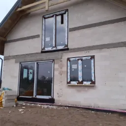 Surowy stan budynku w trakcie montażu stolarki okiennej: widoczne trzy okna z folią ochronną na ramach, ściany z betonu komórkowego i materiały budowlane obok.