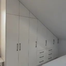 Biała szafa wnękowa z czarnymi uchwytami w pokoju na poddaszu, dopasowana do skosu dachu.