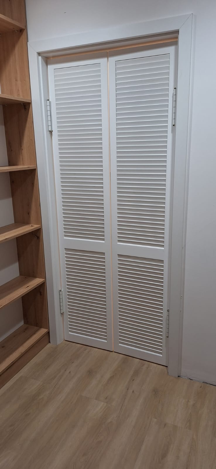 Białe, dwuskrzydłowe drzwi żaluzjowe w garderobie z drewnianymi półkami, widoczne zawiasy i podłoga z paneli.